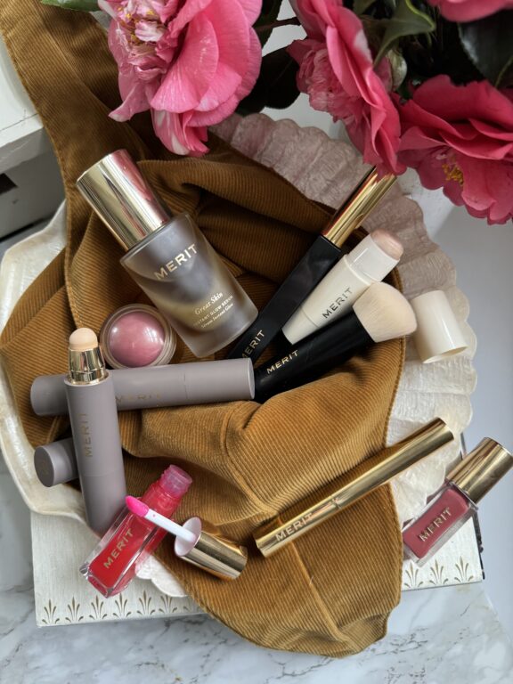 A flat lay of MERIT beauty products, including foundation, blush, brushes, lip gloss, and makeup sticks, arranged on a gold fabric with pink flowers in the corner.