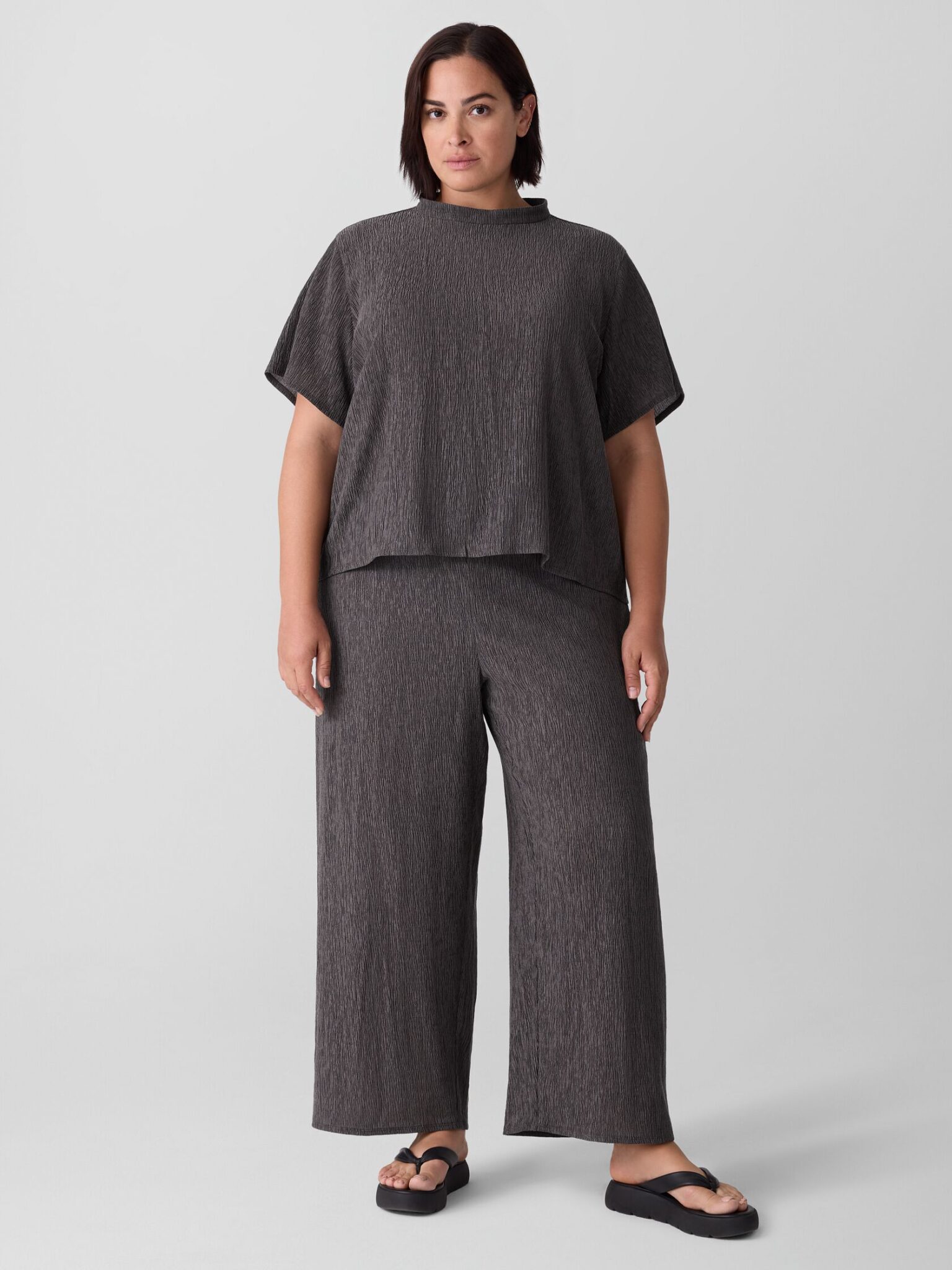 A person stands facing forward, wearing a loose-fitting, dark gray textured top and matching wide-leg pants with black sandals, against a plain light background.