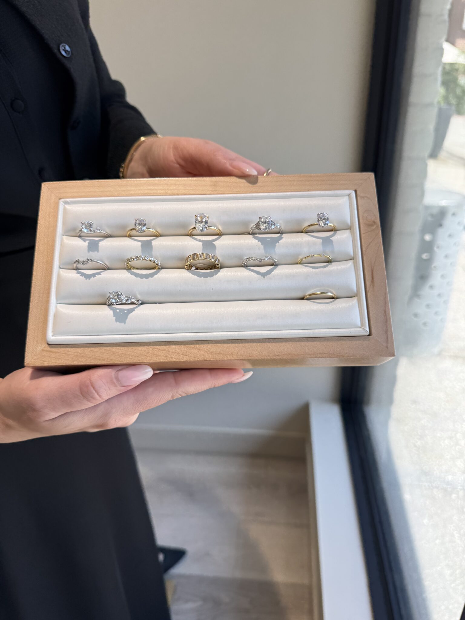 A person holds a wooden display box containing twelve assorted gold and diamond rings, positioned near a window inside a jewelry store.