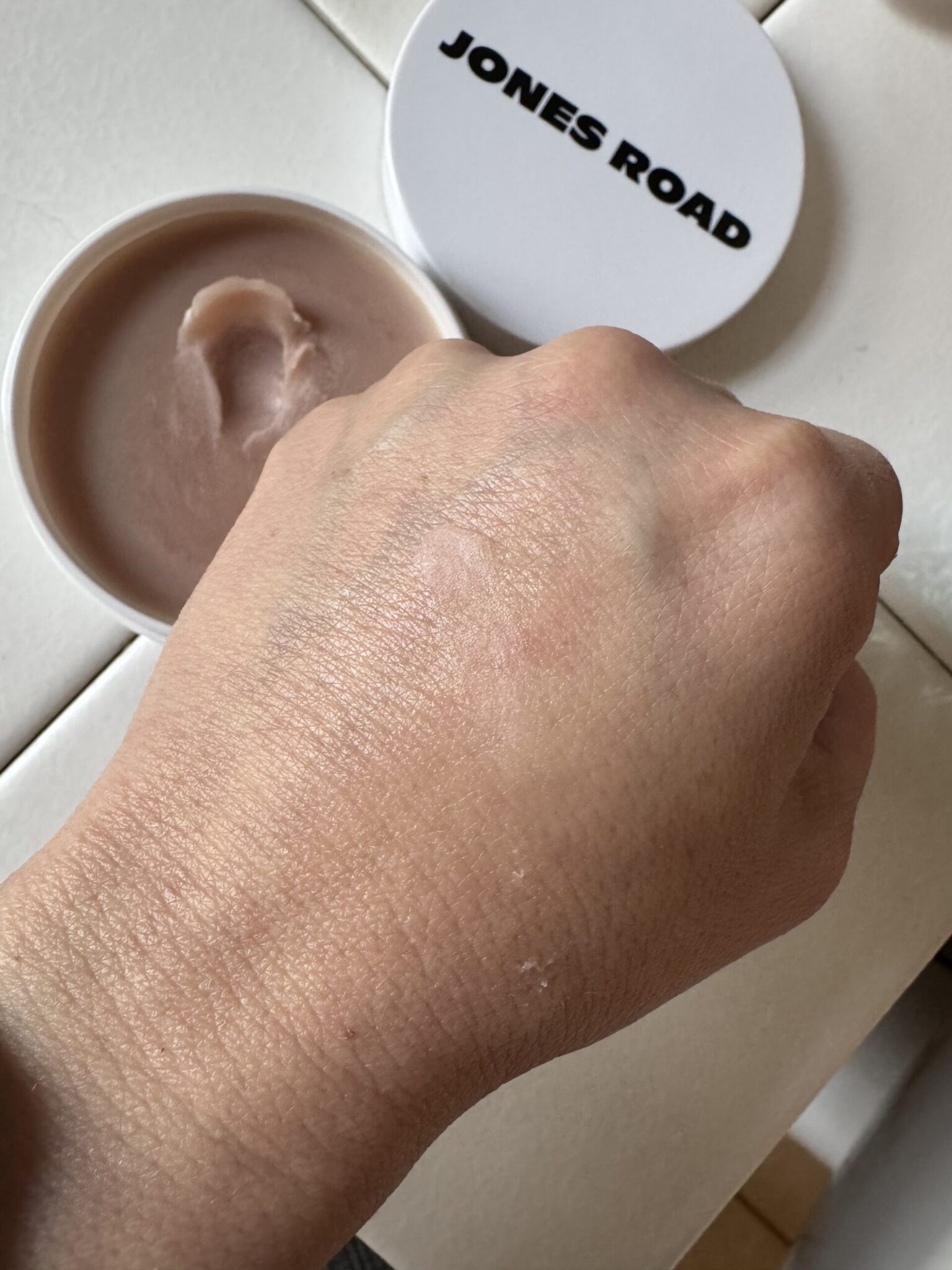 A hand with Jones Road balm applied on the back is shown next to an open container labeled "JONES ROAD." The balm appears lightly spread on the skin.