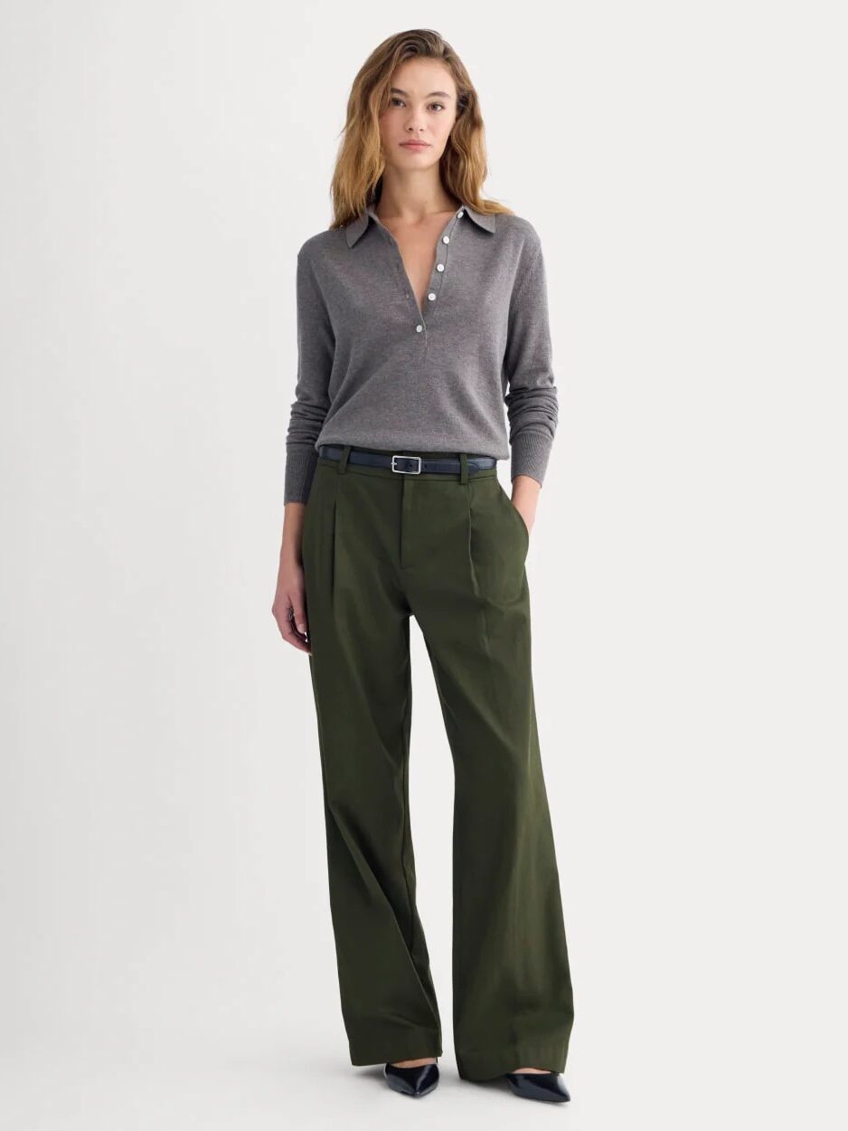A woman stands wearing a grey long-sleeve collared shirt with buttons, olive green wide-leg trousers, a black belt, and black pointed shoes against a plain background.
