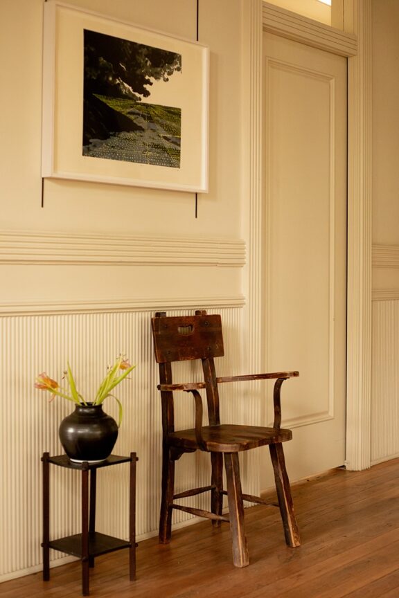 A wooden chair sits against a cream paneled wall below a framed artwork, with a black vase of flowers on a small table nearby.