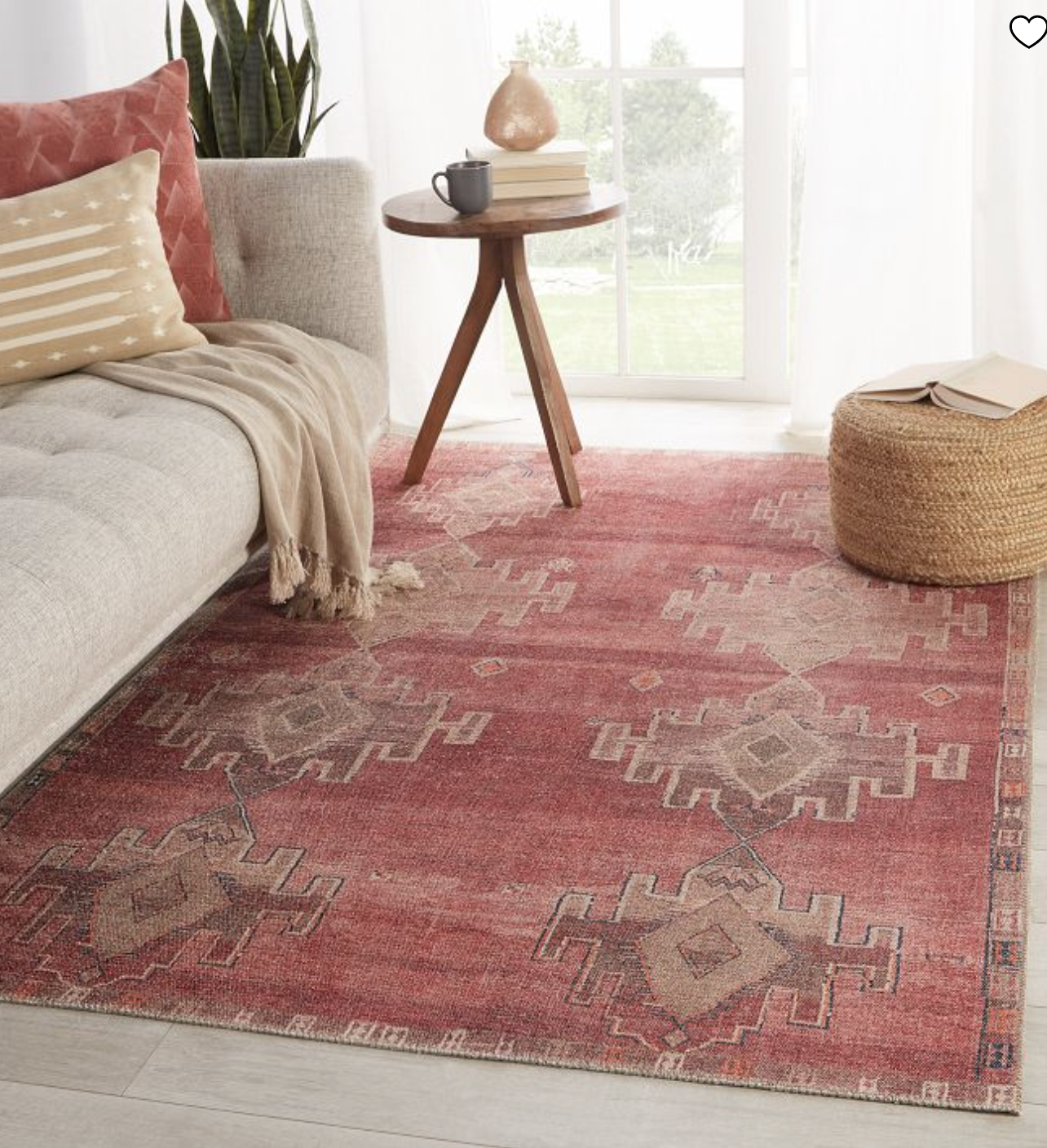A red patterned rug on a light floor near a beige sofa with a striped pillow. A round pouf and a wooden table with a mug and lamp are nearby, with a window view in the background.