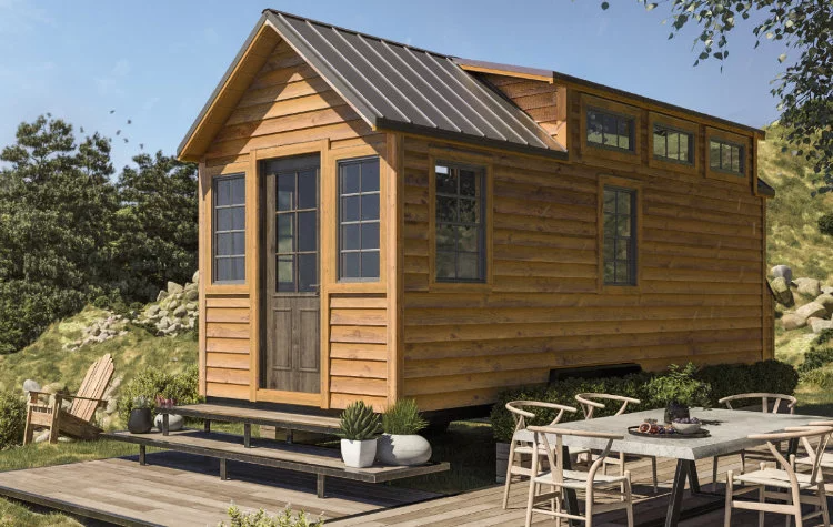A small wooden cabin with a dark metal roof on a platform surrounded by outdoor furniture and plants, located in a grassy area with trees and rocks in the background.